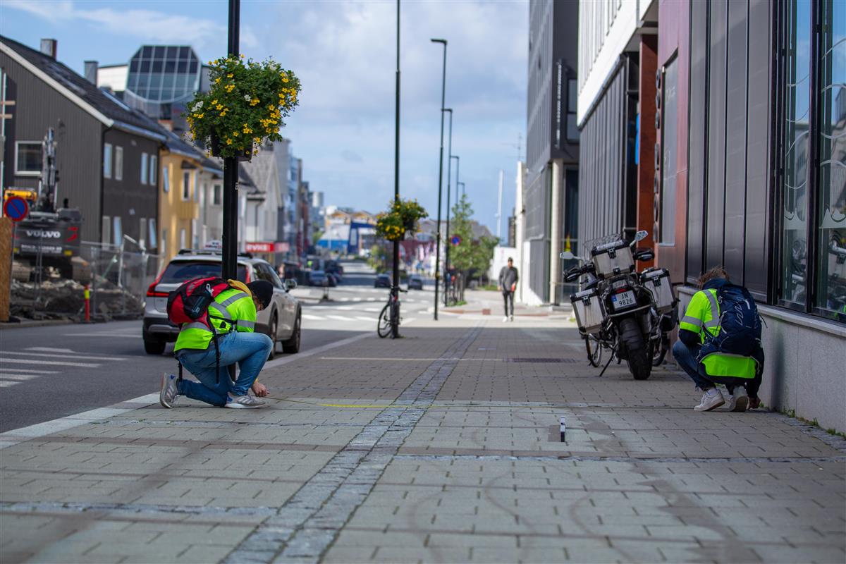 Bildet er tatt av en gang- og sykkelvei i Bodø sentrum. På hver side av fortauet sitter en person på kne. De to personene holder et målebånd mellom seg, som de ser på.  - Klikk for stort bilde