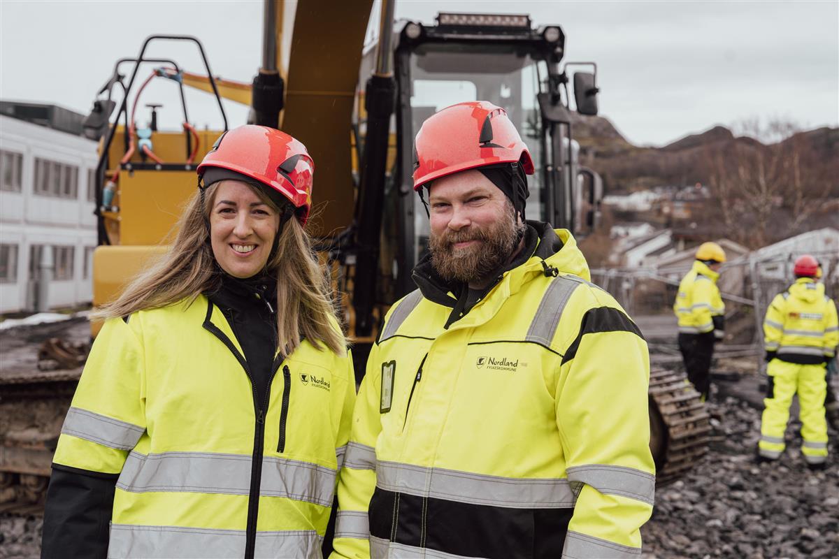 En dame og en mann i anleggstøy foran gravemaskin - Klikk for stort bilde